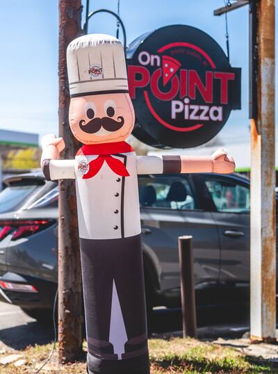 The inflatable chef out front with arms wide beside the On Point Pizza sign