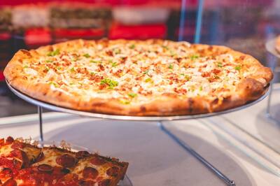 A Chicken Bacon Ranch pizza with chicken, bacon and chopped scallions on a stand in the slice case