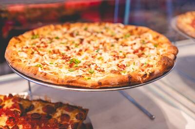 A Chicken Bacon Ranch pizza with chicken, bacon and scallions on a stand in the slice case, with Detroit style pizza below