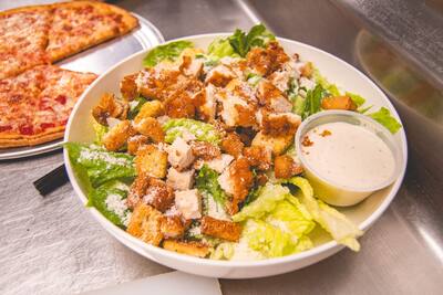 A chicken Caesar salad with croutons and a cup of dressing beside a whole pizza