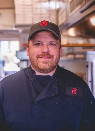 A close portrait of a cook in a cap and black chef coat, smiling