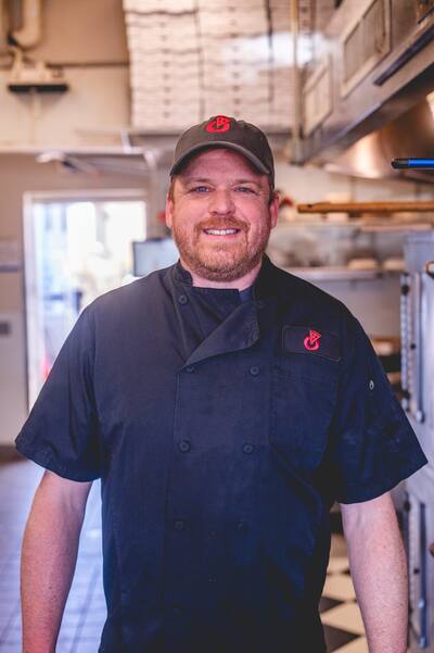 A cook in a black chef coat and cap smiling in the kitchen