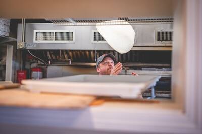 A cook tossing pizza dough in the air, seen through the kitchen window