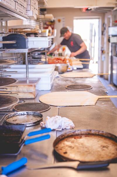 The make line in the kitchen with peels and pans and a cook working at the far end