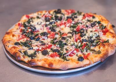 A Mediterranean pizza with spinach, black olives, red onion, tomato and feta on a round tray