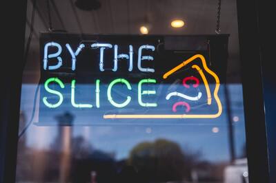 A neon sign in the window that says By the slice