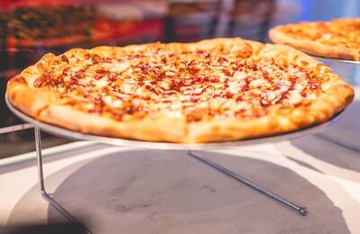 A round pizza with a drizzle of sauce on a stand in the slice case, another pie behind it