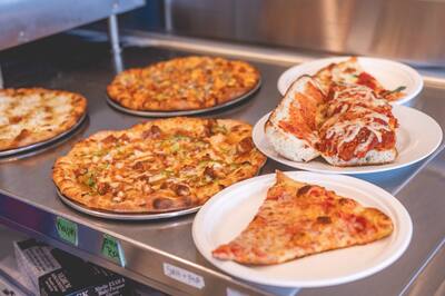 A cheese slice, a sandwich and whole pizzas set out on the counter, from a lower angle